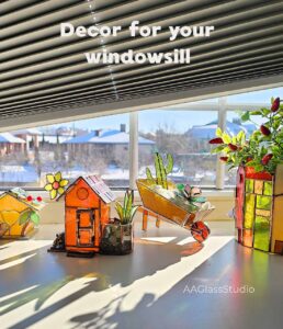 A collection of 3d stained glass pieces on a windowsill: a garden wheelbarrow with succulents, a mini birdhouse, a garden cottage, and a house-shaped flower pot holder, all glowing in natural light.