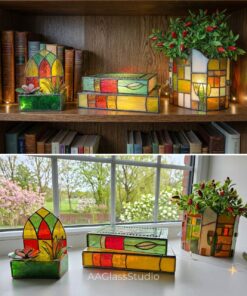 Three 3d stained glass pieces displayed on a windowsill: a mini window planter with succulents, a stained glass stacked books lamp, and a house-shaped planter for a flower pot, all glowing in natural sunlight.