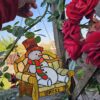 Boss Snowman stained glass suncatcher - hanging on a wooden trellis next to red roses, backlit by the sun